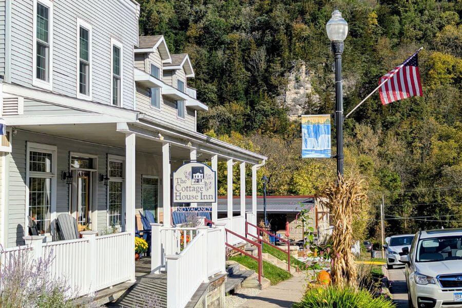 The Cottage House Inn in downtown Lanesboro, Minnesota, decorated with fall touches including cornstalks, pumpkins, and flowers along the sidewalk, with bluffs covered in autumn trees in the background.