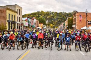 Hundreds of cyclists line up on Lanesboro’s historic downtown street for the start of the Filthy 50 Gravel Bike Race, surrounded by colorful storefronts and autumn bluff scenery.