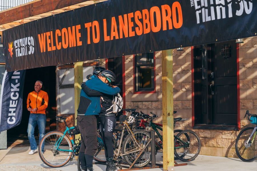 Two cyclists embrace beneath a “Welcome to Lanesboro” Filthy 50 banner outside Sylvan Brewing, surrounded by bikes and fellow riders preparing for the race.