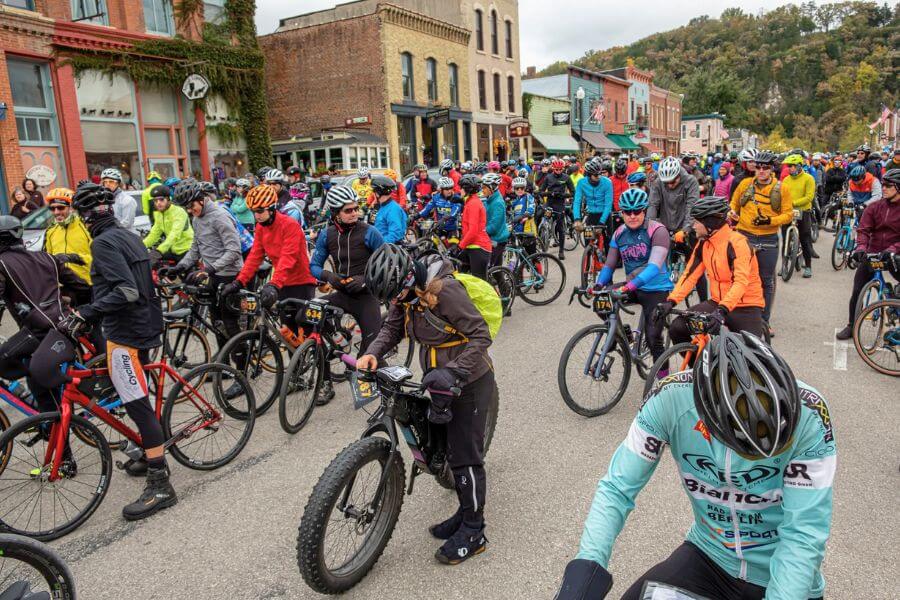 Cyclists gather in colorful gear at the starting line of the Filthy 50 Gravel Bike Race in downtown Lanesboro, surrounded by historic buildings and fall scenery.