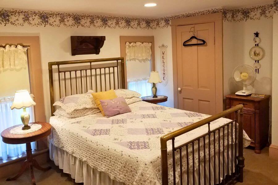 A cozy guest room at the James A. Thompson House Bed & Breakfast in Lanesboro, Minnesota, featuring a vintage brass bed with a handmade quilt, lace-curtained windows, soft lighting from antique lamps, and floral accents that create a warm, welcoming atmosphere.