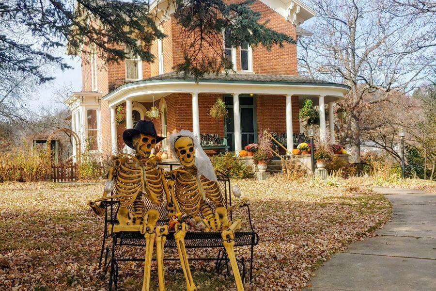 Two Halloween skeleton decorations dressed as a bride and groom sit on a bench in front of the James A. Thompson House Bed & Breakfast in Lanesboro, Minnesota. The historic red-brick Italianate home with white columns is surrounded by autumn leaves, pumpkins, and seasonal decor.