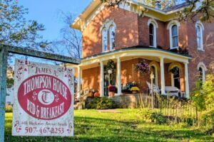 The James A. Thompson House Bed & Breakfast in Lanesboro, Minnesota—a beautifully restored 1870s Italianate brick mansion with white columns, arched windows, and a welcoming front porch decorated with pumpkins and fall flowers. A vintage-style sign in the foreground reads “James A. Thompson House Bed & Breakfast.”