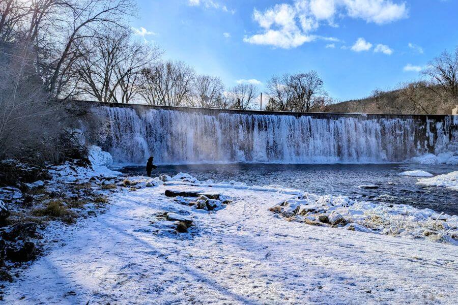 A winter angler stands near the snowy shoreline below the Lanesboro dam, where icy cascades and bright blue skies create a dramatic cold-weather scene.
