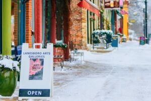 Snow-dusted downtown Lanesboro with colorful storefronts and a sandwich board reading “Lanesboro Arts Galleries – Art Show – Open,” inviting visitors to explore the local gallery on a peaceful winter day.