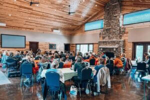 A group gathers inside a rustic wood-paneled event center with high ceilings, large windows, and a stone fireplace, attending a workshop or retreat in Lanesboro during winter.