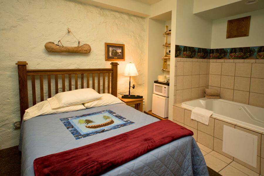 A cozy guest room at the Stone Mill Hotel & Suites in Lanesboro featuring a handcrafted quilt, wooden bed frame, soft lighting, and an in-room whirlpool tub — perfect for a relaxing winter stay.