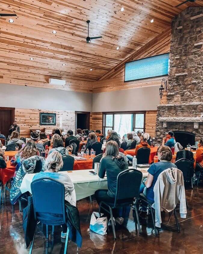 A group gathers inside a rustic wood-paneled event center with high ceilings, large windows, and a stone fireplace, attending a workshop or retreat in Lanesboro during winter.