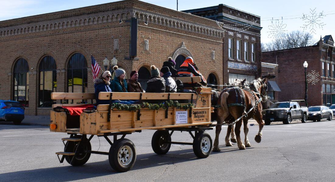 Horse-drawn wagon rides through downtown Lanesboro, Minnesota, with bundled-up visitors enjoying a sunny late-fall day surrounded by historic brick buildings decorated with holiday garlands and lights.