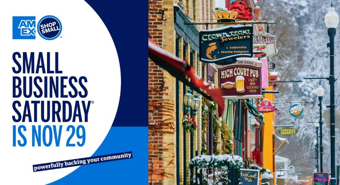 A promotional image for Small Business Saturday featuring the American Express Shop Small logo alongside a snowy winter street scene in downtown Lanesboro, Minnesota, with colorful storefronts and festive decorations.