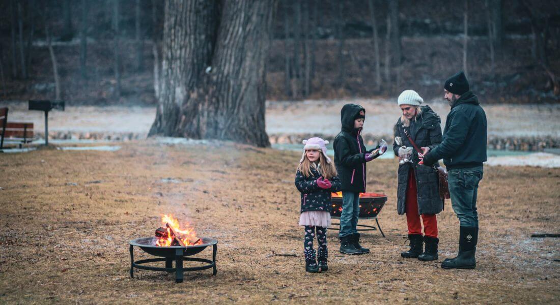 A family gathers around a small outdoor fire pit on a chilly winter evening, preparing s’mores together in a park setting.