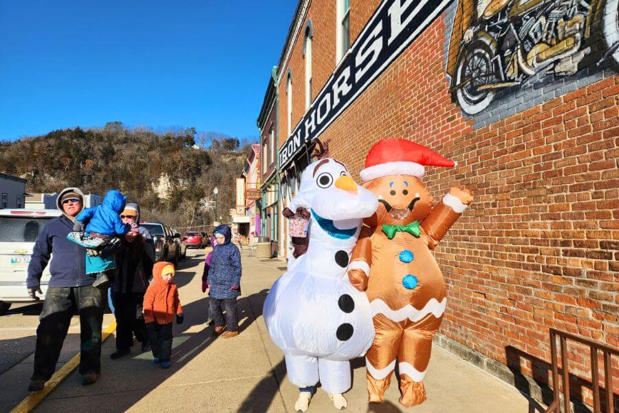 People in winter coats walk along downtown Lanesboro while two costumed characters — a giant gingerbread person in a Santa hat and a snowman — wave on the sidewalk in front of the Iron Horse building.