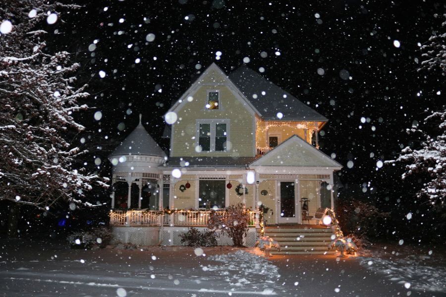 A warmly lit Victorian-style inn in Lanesboro, Minnesota, glows with holiday lights as snow gently falls at night, creating a peaceful and cozy winter scene.