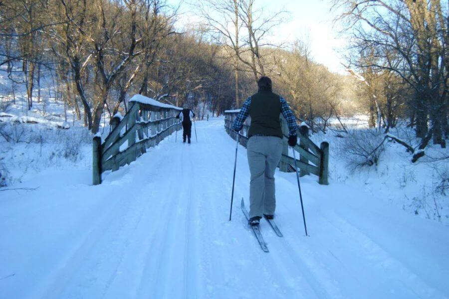 A cross-country skier glides across a snowy bridge on the Root River State Trail on a bright winter day, with another skier in the distance.