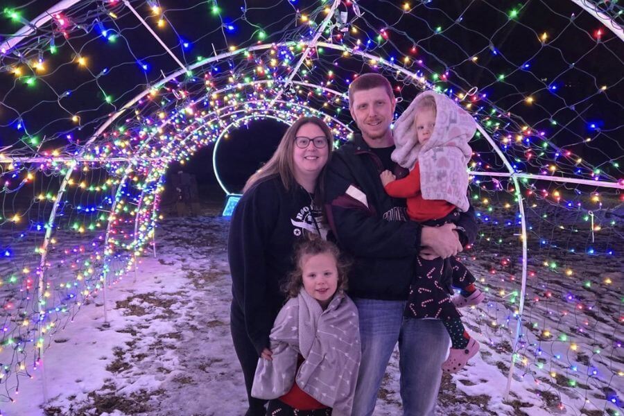 A family stands together beneath a colorful tunnel of holiday lights at the Lanesboro Legion Lights display on a winter night.