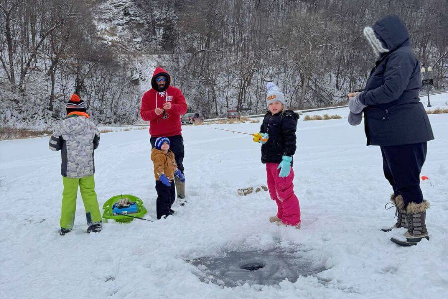 A family bundled up in winter gear participates in ice fishing on a snowy pond, with children holding small fishing rods near a freshly drilled ice hole.