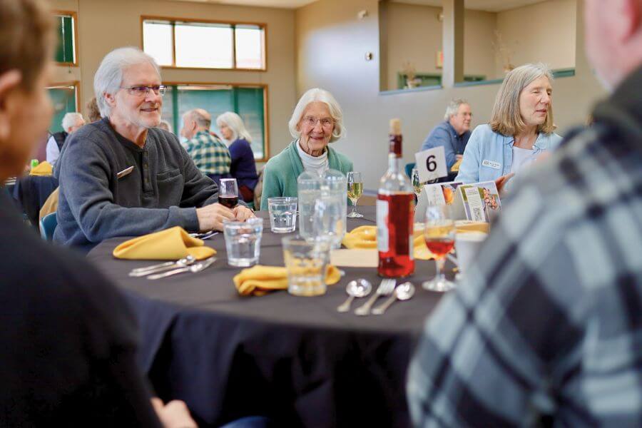 A group of smiling people enjoy food and conversation around a set dining table at a winter gathering inside a bright, spacious event hall in Lanesboro, Minnesota.