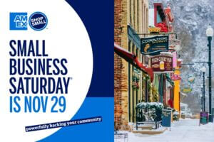 A promotional image for Small Business Saturday featuring the American Express Shop Small logo alongside a snowy winter street scene in downtown Lanesboro, Minnesota, with colorful storefronts and festive decorations.