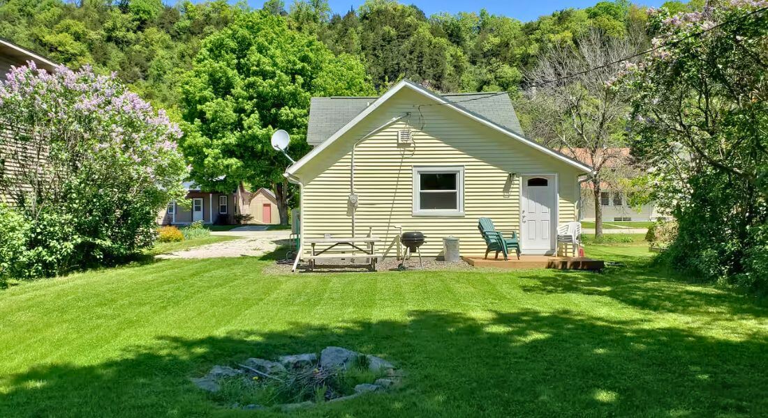 Backyard of The Cozy Cottage in Lanesboro, MN, with green lawn, fire pit, grill, and seating set against Bluff Country hills.