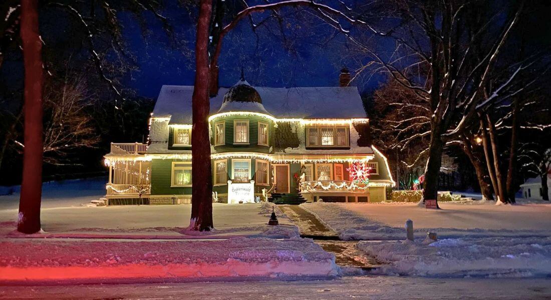 Historic Oakenwald Terrace mansion in Chatfield, Minnesota illuminated with warm white Christmas lights on a snowy winter evening, showcasing its Victorian architecture, wraparound porch, and festive holiday decorations under a dark blue sky.