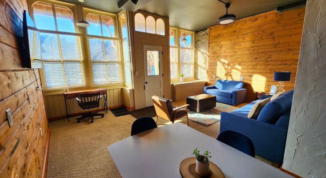 A bright, inviting sitting room with large sun-filled windows, warm wood-paneled walls, and cozy blue furniture at Root River Inn & Suites, featuring a workspace, lounge seating, and a dining table—an ideal setting for relaxed productivity during a corporate or group retreat in Lanesboro.