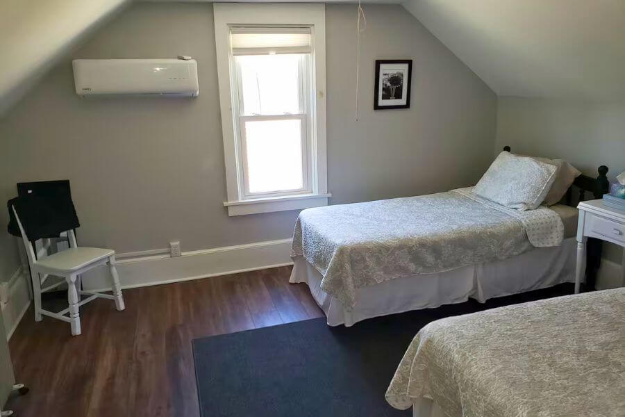 Cozy bedroom at The Cozy Cottage in Lanesboro, MN, with two twin beds, sloped ceiling, window light, and mini-split air conditioning.
