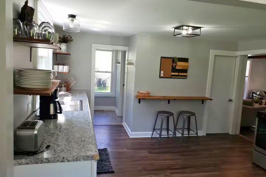Another view of the bright kitchen at The Cozy Cottage in Lanesboro, MN, with open shelving, coffee station, granite counters, and a breakfast bar with stools.