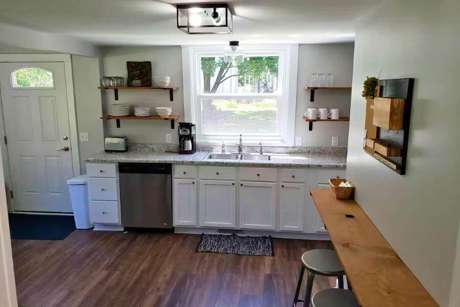 Bright, fully equipped kitchen at The Cozy Cottage in Lanesboro, MN, with white cabinets, open shelving, dishwasher, coffee maker, and breakfast bar.