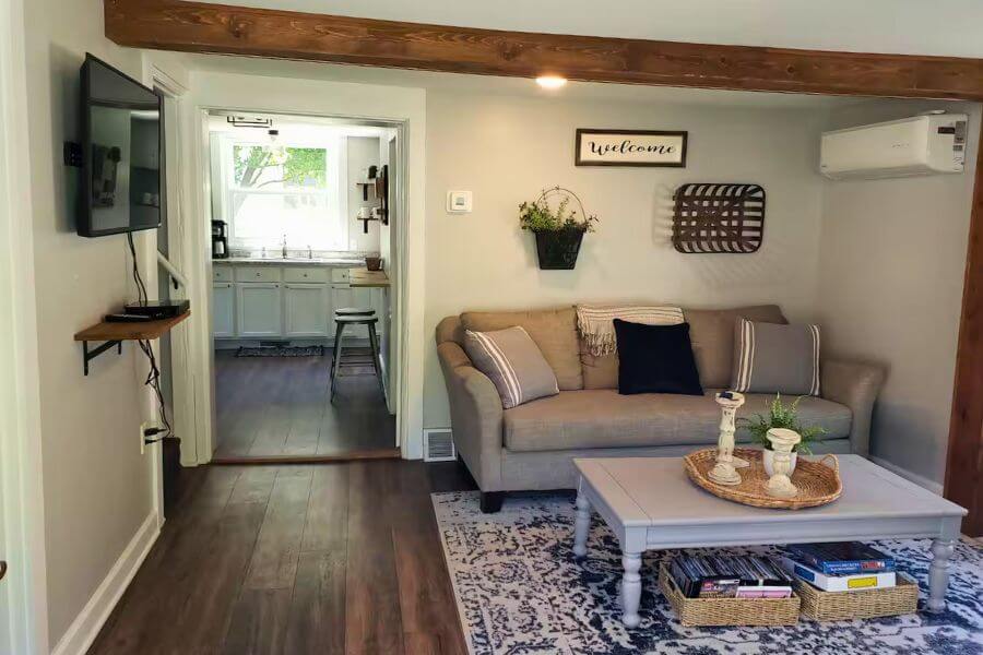 Living room at The Cozy Cottage in Lanesboro, MN, with cozy sofa, coffee table, Smart TV, wood beam accents, and kitchen visible in the background.