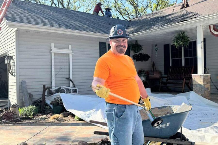 A smiling construction worker in a hard hat and gloves stands in front of a home with roofing work underway, holding a shovel beside a wheelbarrow during a residential project.