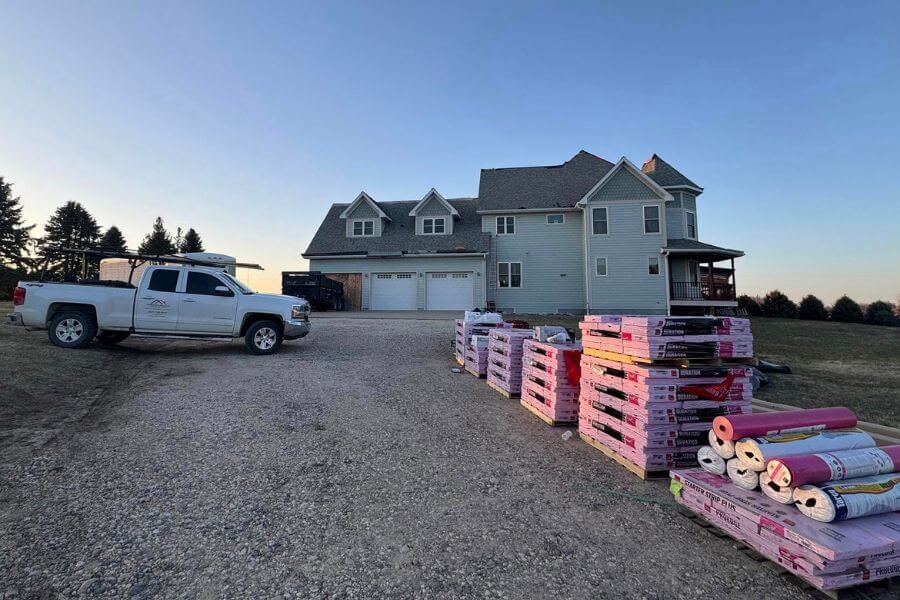Stacks of roofing shingles and materials sit in front of a large home as a contractor truck is parked nearby, ready for a roof replacement project in southeast Minnesota.