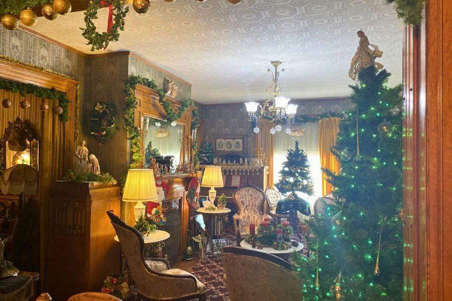 Victorian parlor at Oakenwald Terrace decorated for Christmas, featuring multiple lit Christmas trees, antique furniture, ornate woodwork, garlands, vintage lamps, and a detailed fireplace mantel adorned with holiday décor.