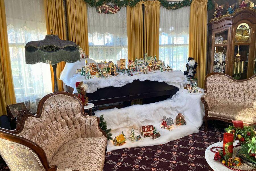 Victorian sitting room at Oakenwald Terrace featuring antique furniture and a holiday village display arranged on top of a piano, surrounded by faux snow, miniature houses, trees, and festive decorations.