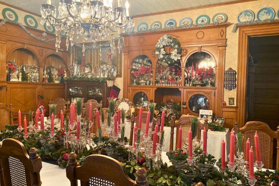 Victorian dining room inside Oakenwald Terrace decorated for Christmas, featuring ornate woodwork, a crystal chandelier, wreaths, poinsettias, and a long table set with pink taper candles, greenery, and antique glassware.