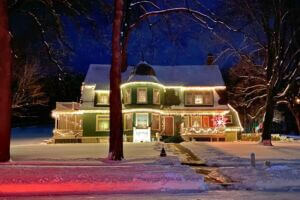 Historic Oakenwald Terrace mansion in Chatfield, Minnesota illuminated with warm white Christmas lights on a snowy winter evening, showcasing its Victorian architecture, wraparound porch, and festive holiday decorations under a dark blue sky.