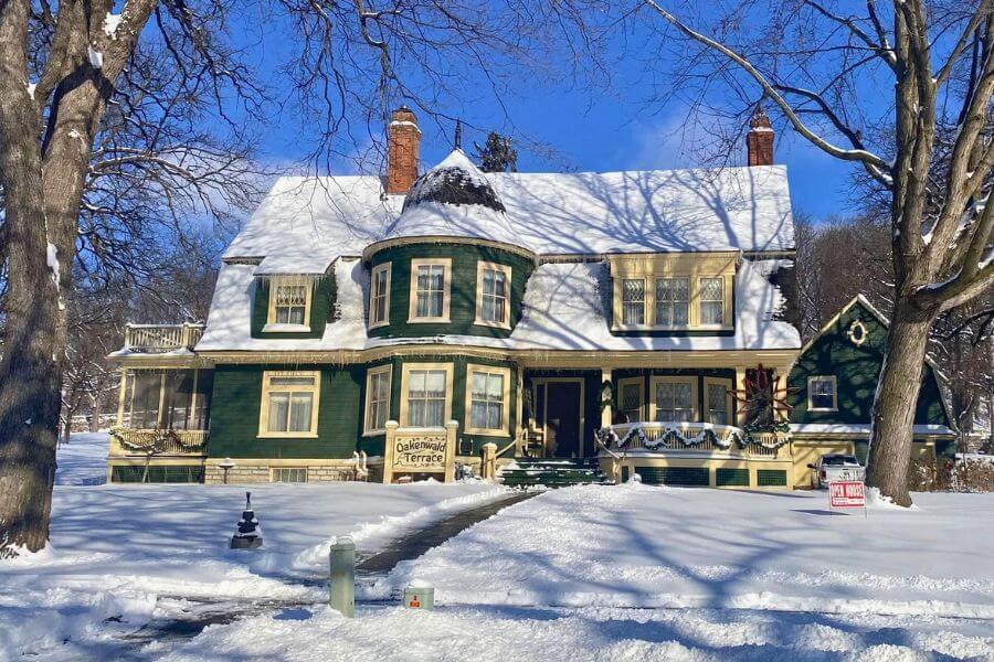 Oakenwald Terrace mansion in Chatfield, Minnesota on a bright winter day, covered in fresh snow. The historic green and cream Victorian home features a turret, screened porch, and holiday decorations along the porch railing.