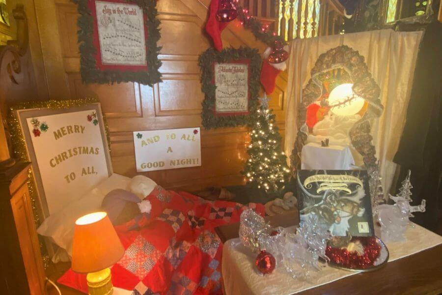 Christmas display inside Oakenwald Terrace featuring a cozy bed with a patchwork quilt, signs reading “Merry Christmas to All” and “And to All a Good Night,” sheet-music décor, stockings, a small lit Christmas tree, and a scene of Santa by a mirror framed in garland.