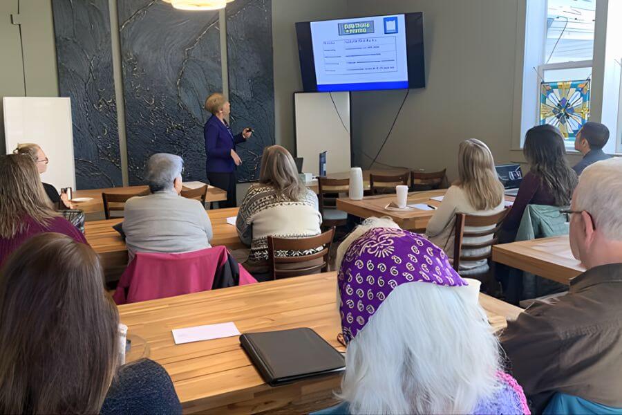 A group of adults seated at wooden tables in the Back Room at Paddle On Coffee in Lanesboro, attentively watching a presenter give a slideshow on a wall-mounted screen during a workshop or corporate training session.