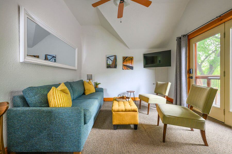 A bright, modern sitting room at Root River Inn & Suites featuring a teal sofa with yellow pillows, mid-century style chairs, a matching ottoman, and large glass doors opening to a tree-filled view—an inviting space for relaxation or team conversations during a Lanesboro retreat.