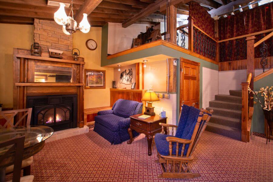 Interior of the Buffalo Bill Suite at Stone Mill Hotel & Suites, featuring a warm gas fireplace with a wooden mantel, cozy upholstered chairs, a lofted sleeping area with rustic railings, exposed beams, and inviting frontier-inspired décor.