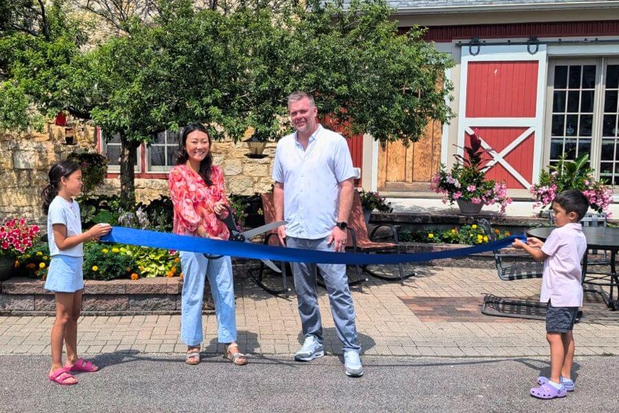Annie Kang and Adam McCabe cut a blue ribbon outside Stone Mill Hotel & Suites during a sunny ribbon-cutting celebration, with two children holding the ribbon ends. Flowers, stonework, and red barn-style doors are visible in the background.