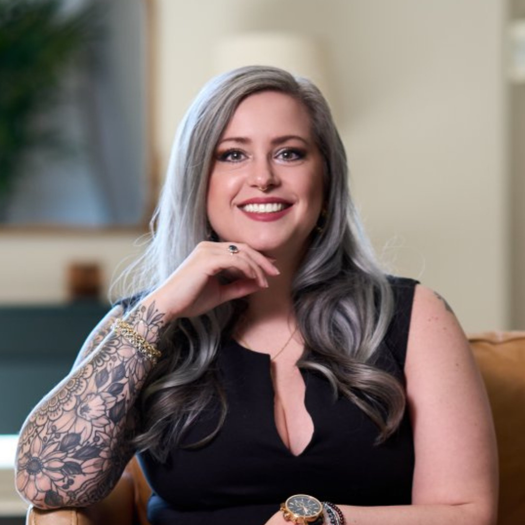 Katherine Frisby Woman sitting in a chair smiling with her hand on her chin with a black top on