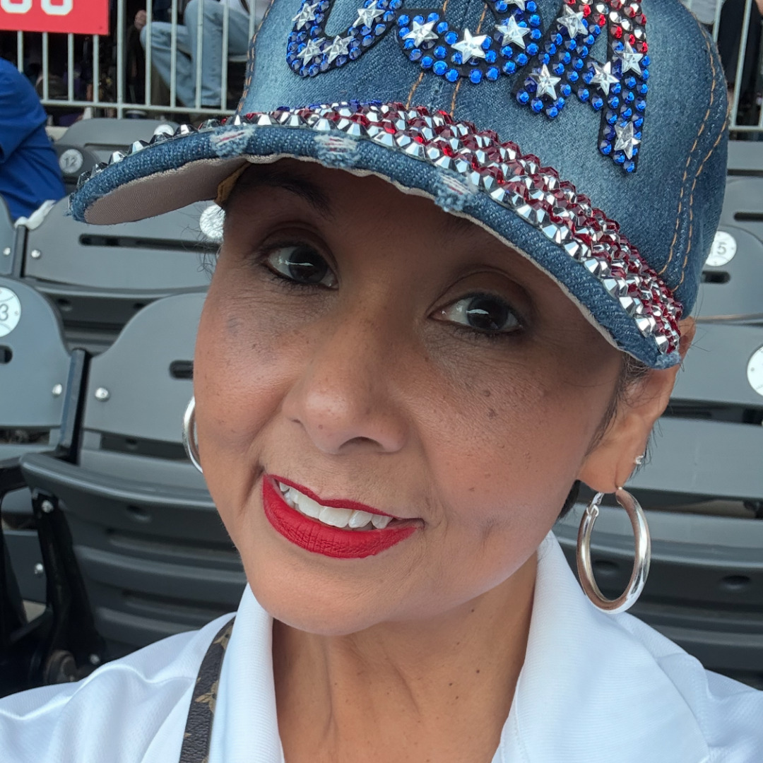 woman smiling in white shirt and denim hat that reads "U.S.A." in the stands of a baseball field