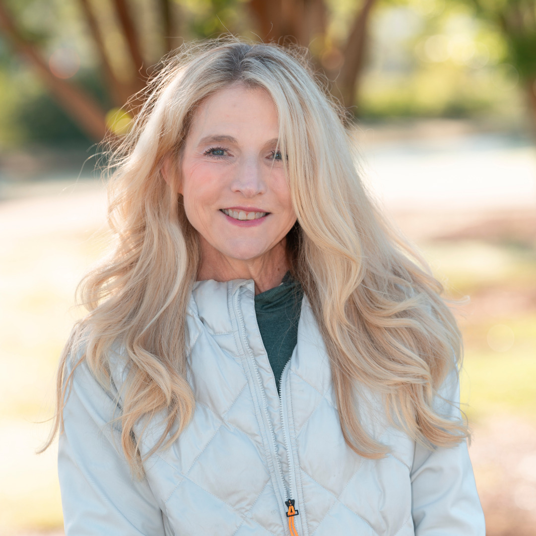 Suzanne Swift woman with blonde hair in a puffy white coat smiling outside against a tree