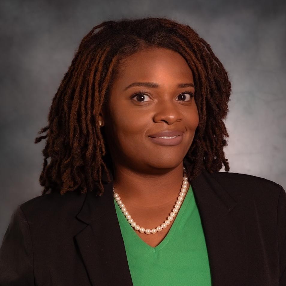 headshot Woman in black blazer and green top with a pearl necklace smiling against a gray background