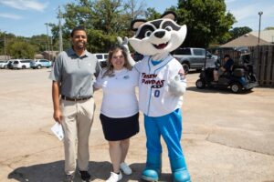 A man and a woman dressed for a golf tournament, standing with a large raccoon mascot of the Rocket City Trash Pandas. They are standing in a parking lot and smiling.