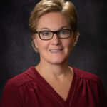 Heidi Huber This is a professional portrait of a woman in a mid‑career age range, likely in her 40s or early 50s. She has short, neatly styled light brown hair with soft waves, and she’s wearing rectangular black‐framed glasses that highlight her warm, light-colored eyes. Her expression is friendly and approachable, with a gentle smile. She’s dressed in a deep burgundy blouse with subtle gathering at the shoulders, and she wears a delicate pendant necklace and small drop earrings. The background is a dark, softly textured backdrop that keeps the focus on her face.