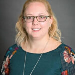 erin karlosky She’s a woman with shoulder‑length wavy blonde hair and rectangular glasses, smiling warmly at the camera. She wears a teal top with floral‑patterned sleeves and a long silver necklace against a neutral gray backdrop.