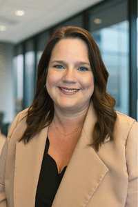 Photo of a smiling woman named Megan Gross wearing a tan blazer and black blouse, standing in front of a wall of windows while looking directly into the camera.