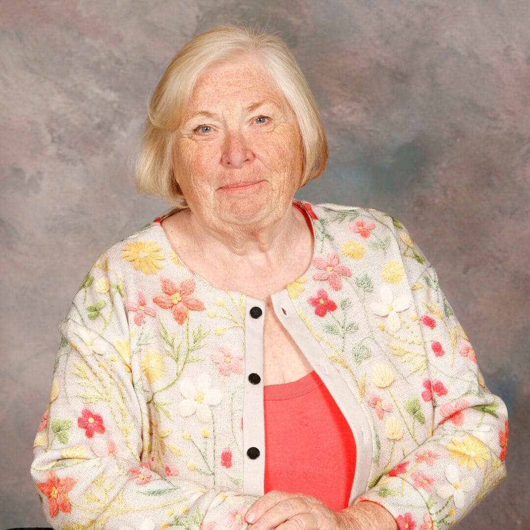 Niki Carlan, Vice-President Advocacy Professional portrait of Niki Carlan seated with hands folded, wearing a floral-patterned jacket over a coral top against a mottled grey background.
